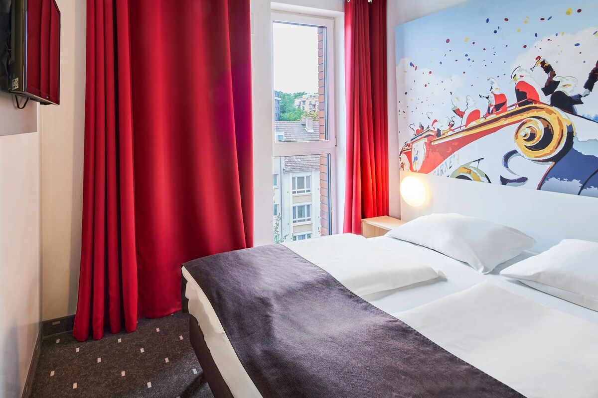 Quarto de hotel com uma cama, cortinas vermelhas, janela com vista para edifícios e uma parede com um mural colorido.