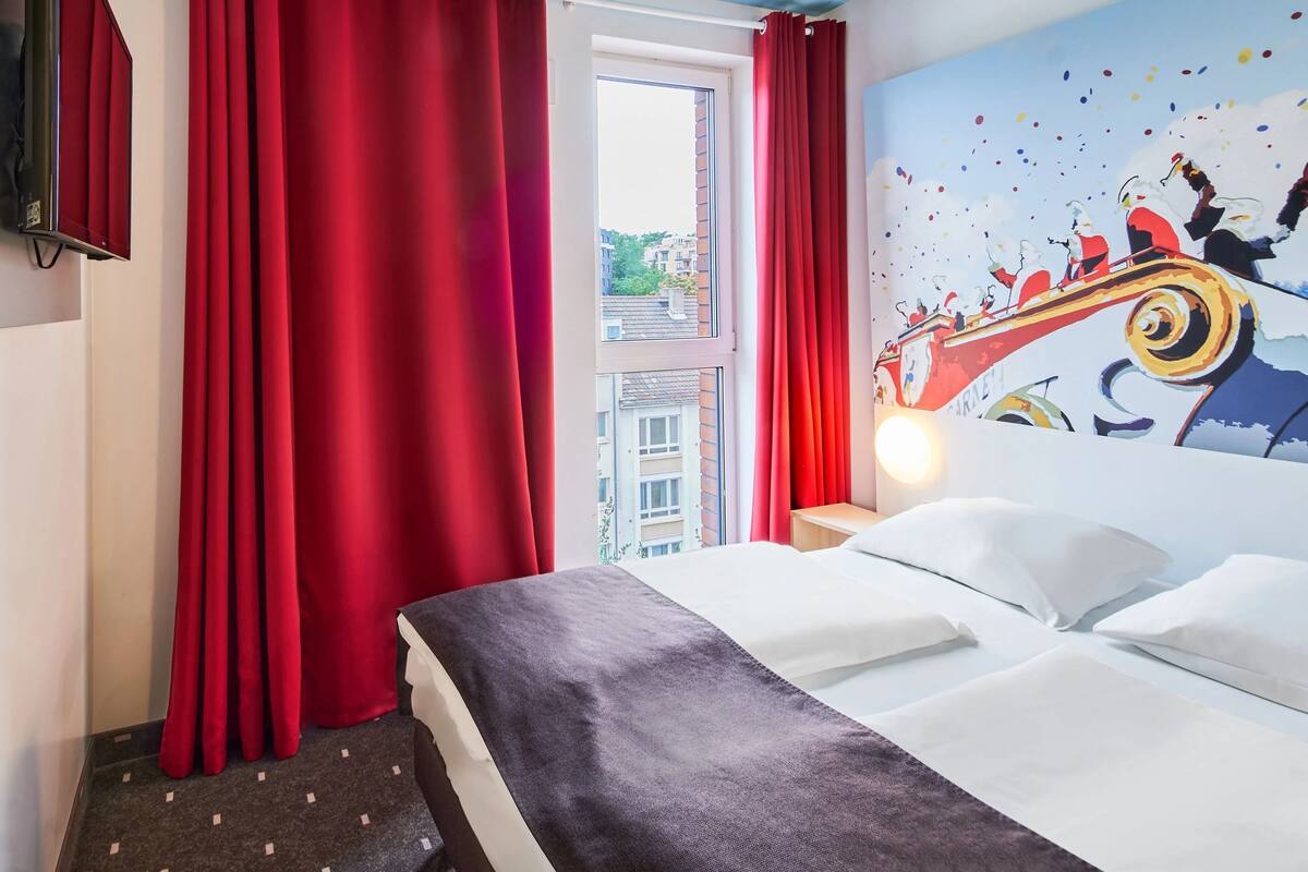 Ein Hotelzimmer mit Bett, roten Vorhängen, einem Fenster mit Blick auf Gebäude und einer Wand mit einem farbenfrohen Wandgemälde.