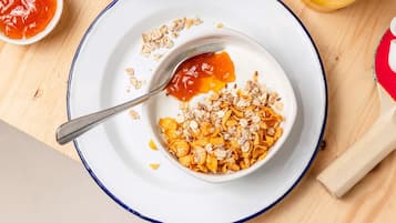 Une table mise pour le petit-déjeuner, avec un bol de céréales, une cuillère et un petit bol de confiture sur une assiette blanche à bordure bleue.