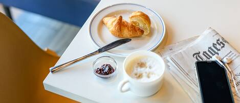 A table with a croissant, a cup of coffee, a knife, a small bowl of jam, and a newspaper with a smartphone on it.