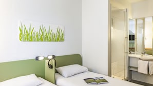 A hotel room with a bed, bedside lamps, a green headboard, a wall-mounted artwork, and a bathroom with a mirror, sink, and towel rack. - ibis budget Zurich City West (Zürich)