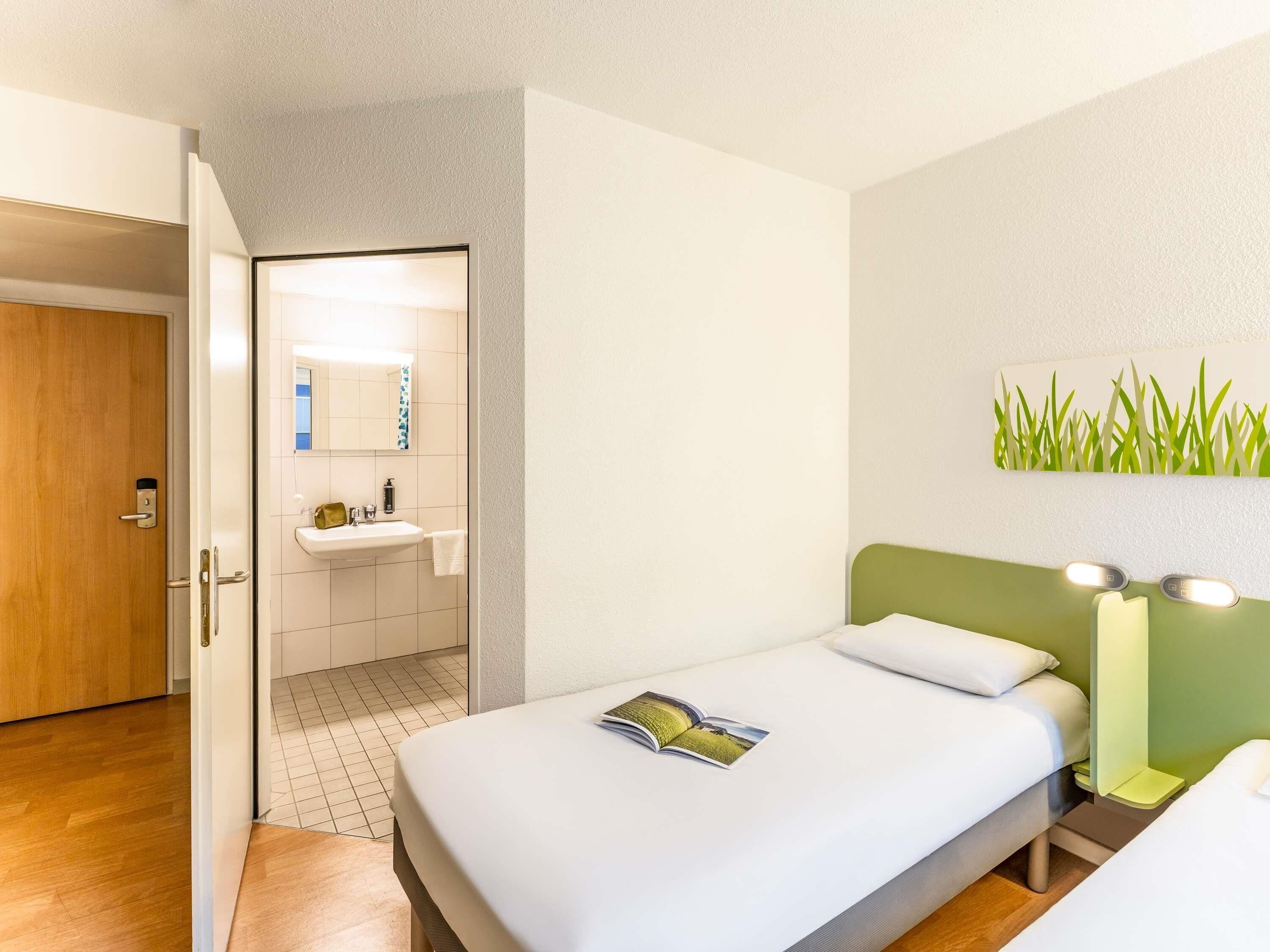 Une chambre d’hôtel avec un lit, une table de chevet, une tête de lit verte, une lampe fixée au mur, une salle de bain avec un lavabo et un miroir, et une porte en bois.