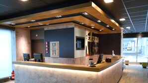 A reception desk with a modern design, featuring a marble countertop and a wooden accent wall. There are fire extinguishers mounted on the wall, and a television is visible above the counter. - ibis budget Zurich City West (Zürich)
