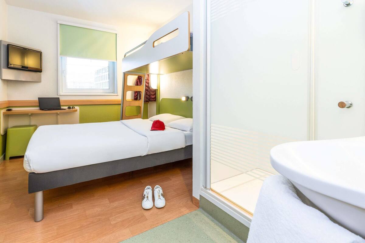 A compact hotel room with a bunk bed, a desk with a laptop, a small seating area, and a bathroom.