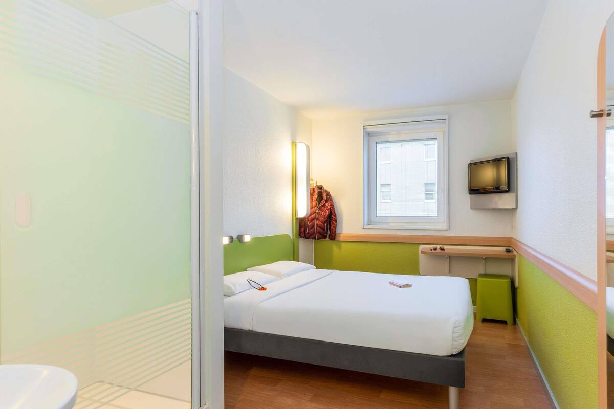 A compact hotel room with a single bed, a small desk, a television, and a window with blinds.
