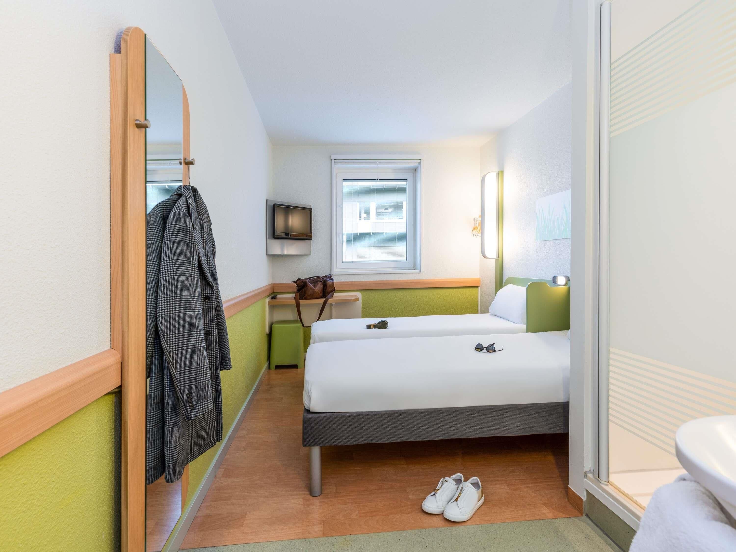 A hotel room with a bed, a mirror, a TV, a window, and a pair of shoes on the floor.