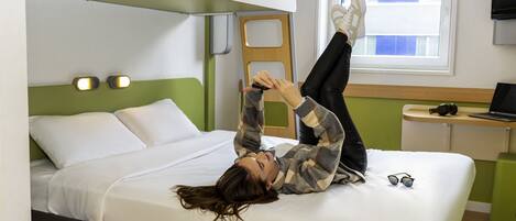 A bed with a green headboard, a window with blinds, and a laptop on a bedside table.