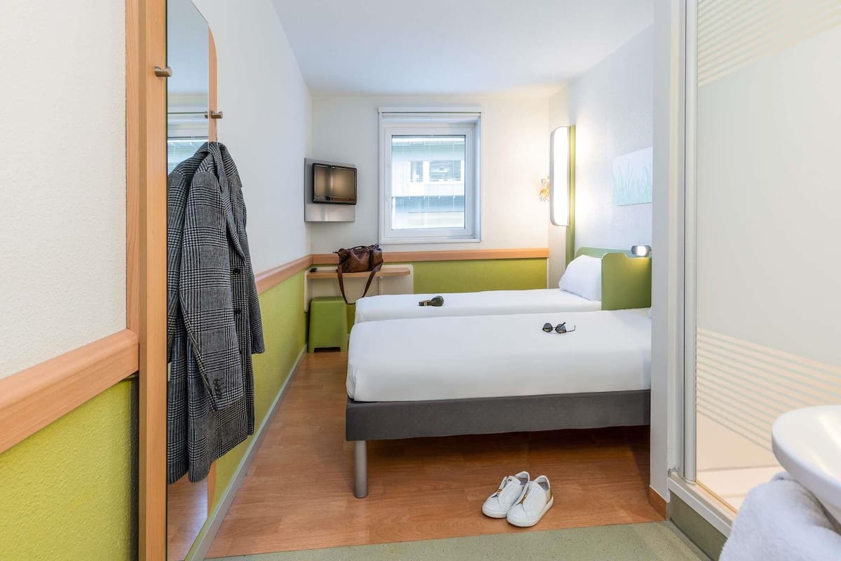 A hotel room with a bed, a mirror, a TV, a window, and a pair of shoes on the floor.