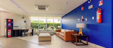 Un vestíbulo moderno con una pared azul decorativa, un extintor, una máquina expendedora y una zona de estar con sofá y sillones.