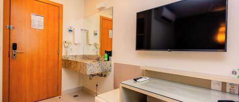 A hotel bathroom with a wooden door, a sink with a mirror, a television mounted on the wall, and a trash bin.