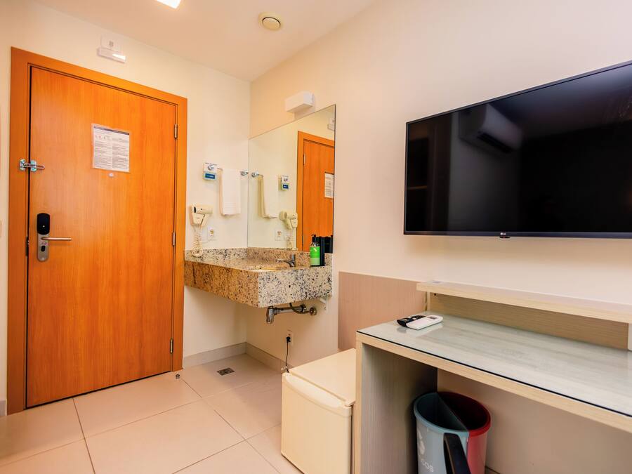 Une salle de bain d’hôtel avec une porte en bois, un lavabo avec un miroir, un téléviseur fixé au mur et une poubelle.