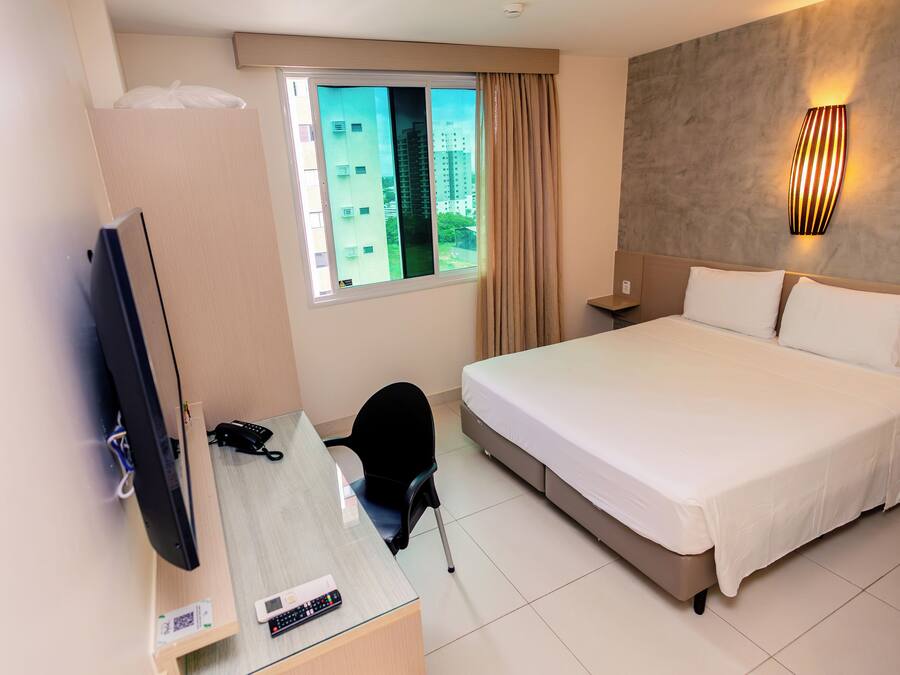 A hotel room with a bed, a desk with a computer, a chair, a TV, and a window with curtains.