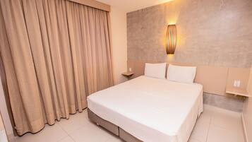 A hotel room with a large bed, beige curtains, a wall-mounted light fixture, and a small bedside table.