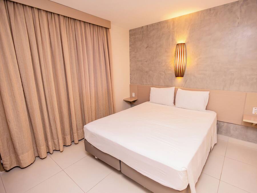 A hotel room with a large bed, beige curtains, a wall-mounted light fixture, and a small bedside table.