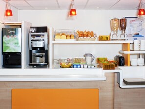 A hotel reception area with a coffee machine, a refrigerator, a fruit bowl, and a buffet with bread and pastries. - ibis budget Orgeval (Orgeval)