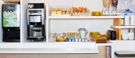 Zona de recepción de un hotel con una máquina de café, un refrigerador, un frutero y un bufé con panadería.