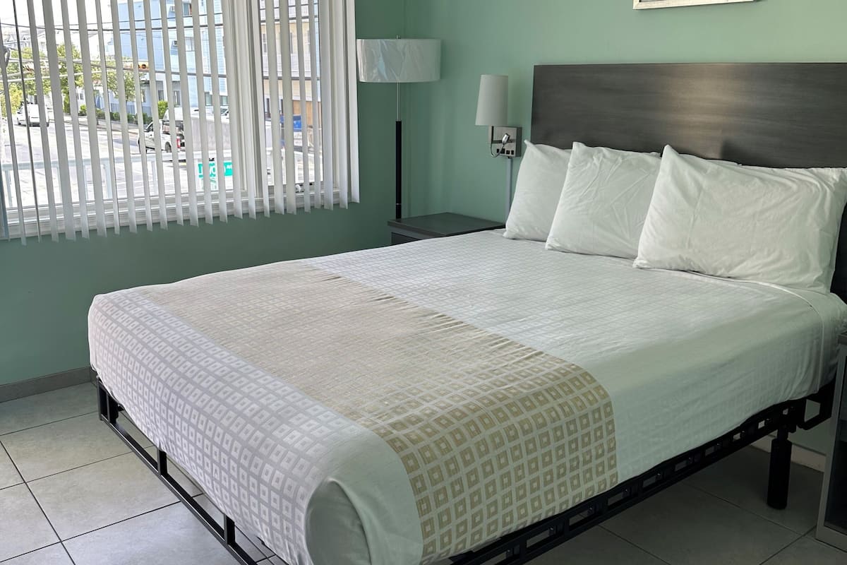 A neatly made bed in a room with a view of the street through the window.