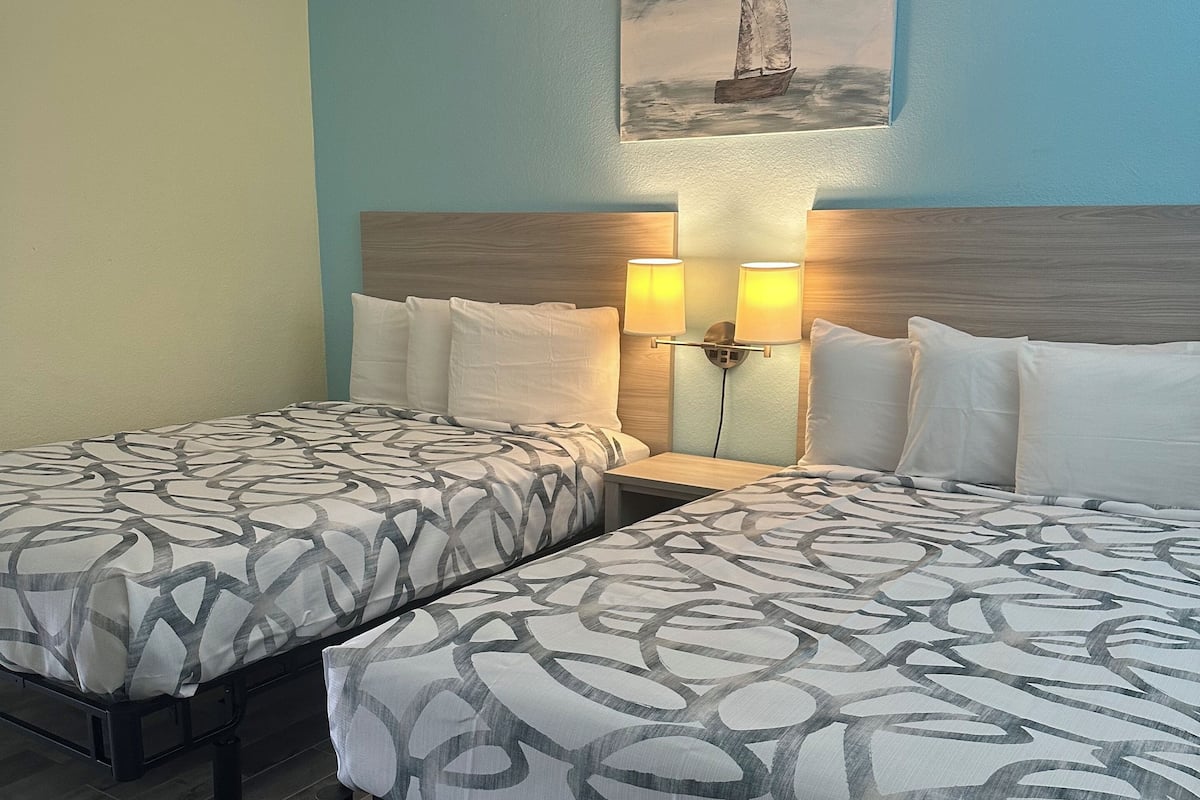 A hotel room with two beds, each with patterned bedding and headboards. There are two wall-mounted lamps on the nightstand between the beds.