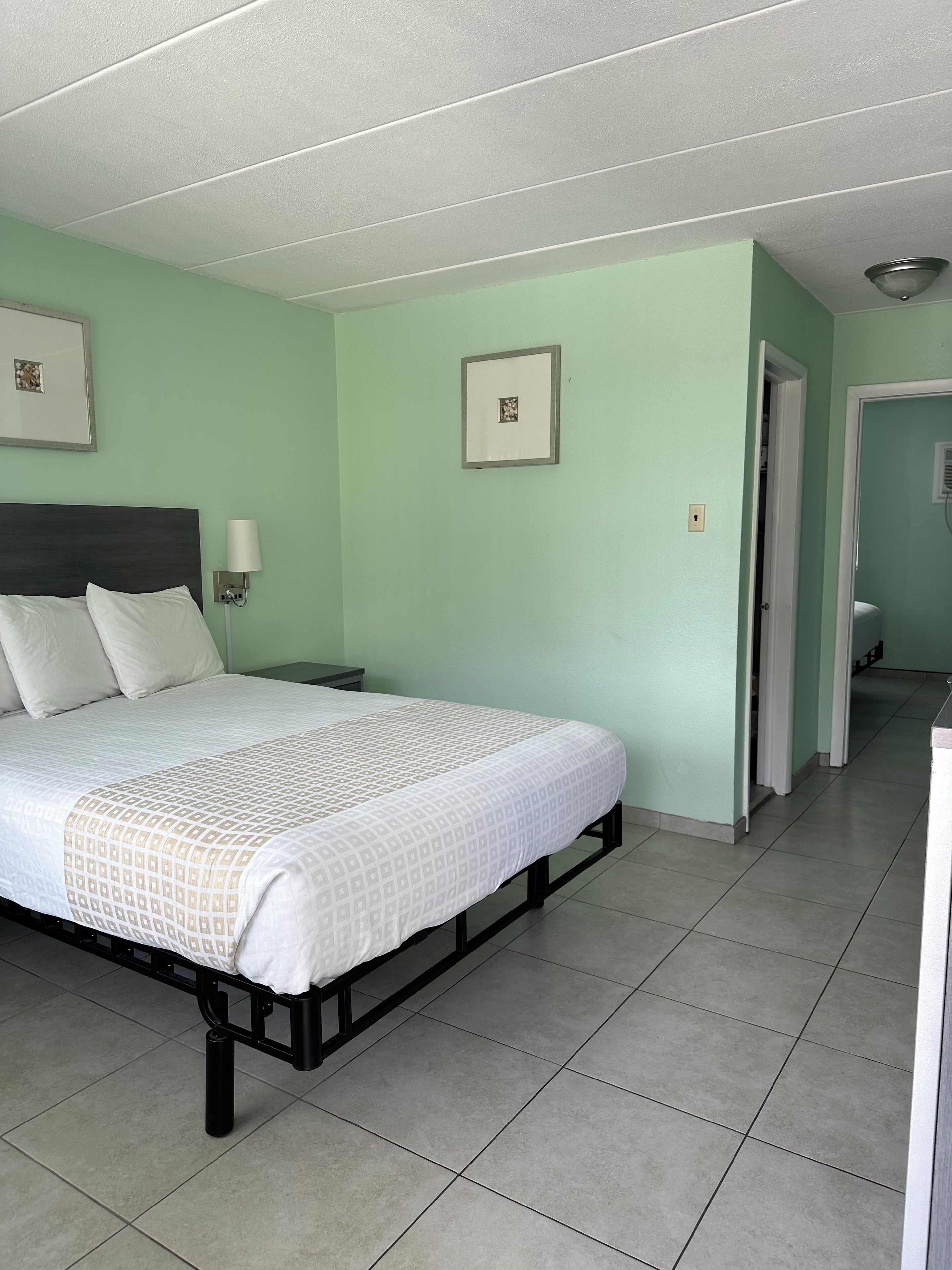 A neatly made bed with a white and beige patterned bedspread, a dark headboard, and a bedside lamp. The room has light green walls, a tiled floor, and a door leading to another room.
