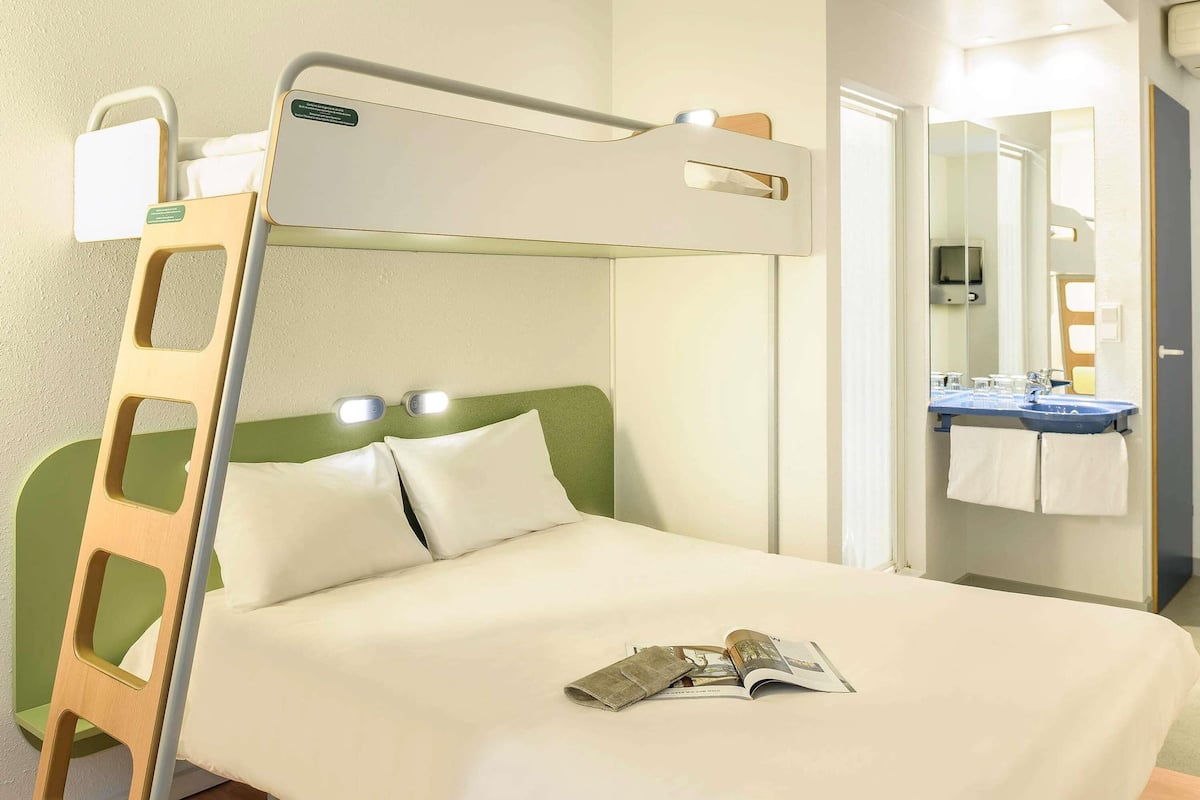 A single bed with a headboard, a bedside table, a lamp, a mirror, and a bathroom with a sink and towel rack.