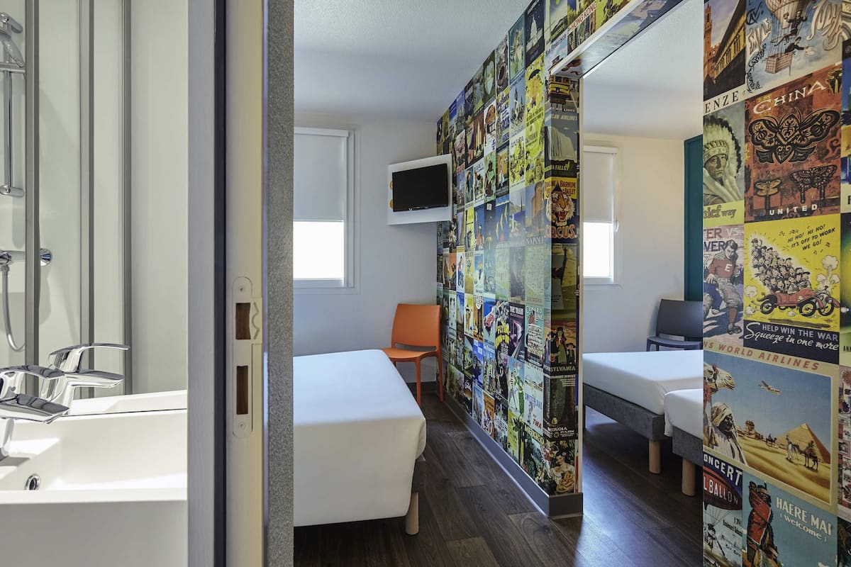 A hotel room with a shower, two beds, a TV, and a wall covered with vintage travel posters.