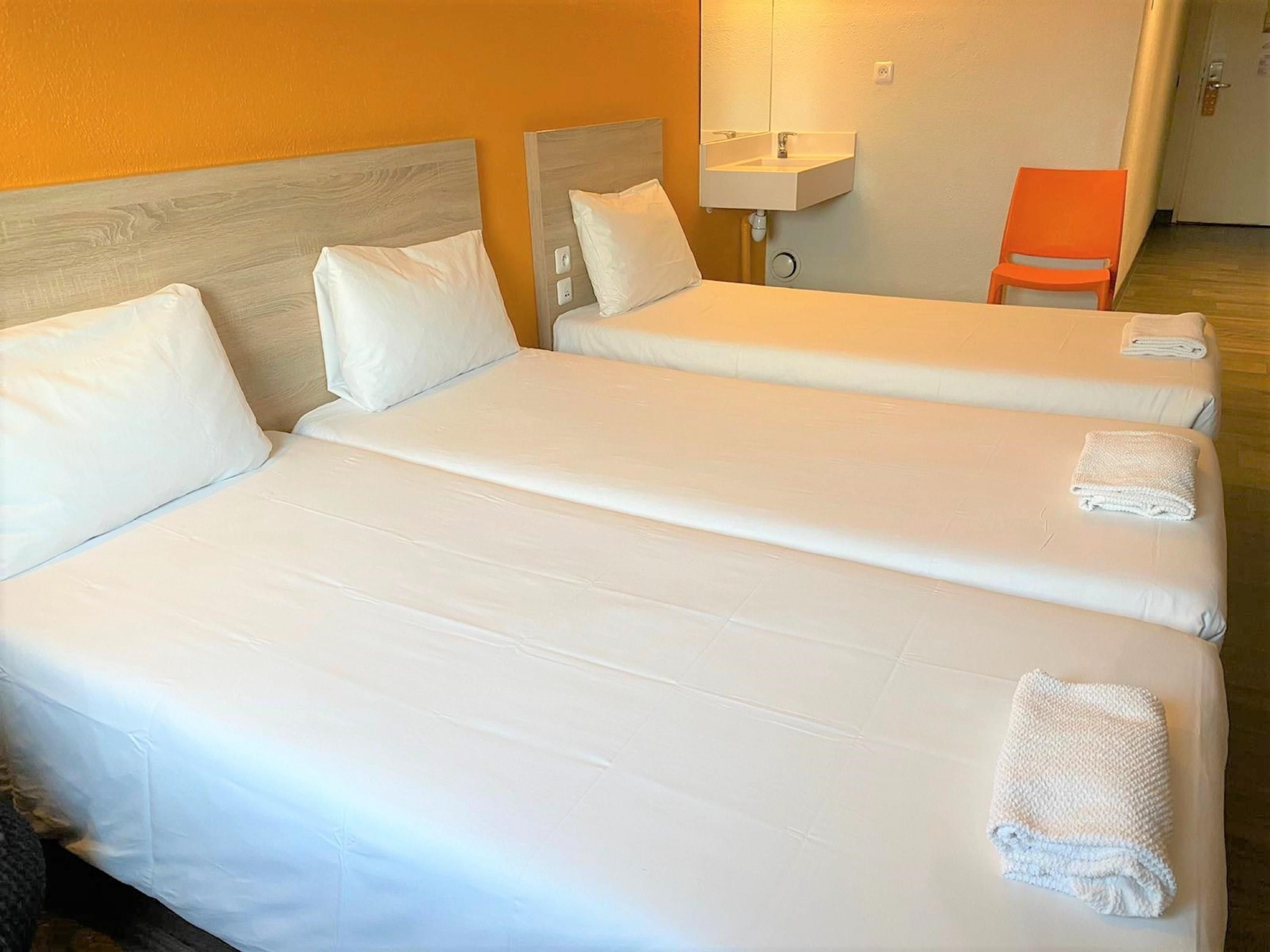 Habitación de hotel con tres camas individuales, cada una con ropa blanca y almohadas, una mesita de noche con lámpara, y una silla naranja.