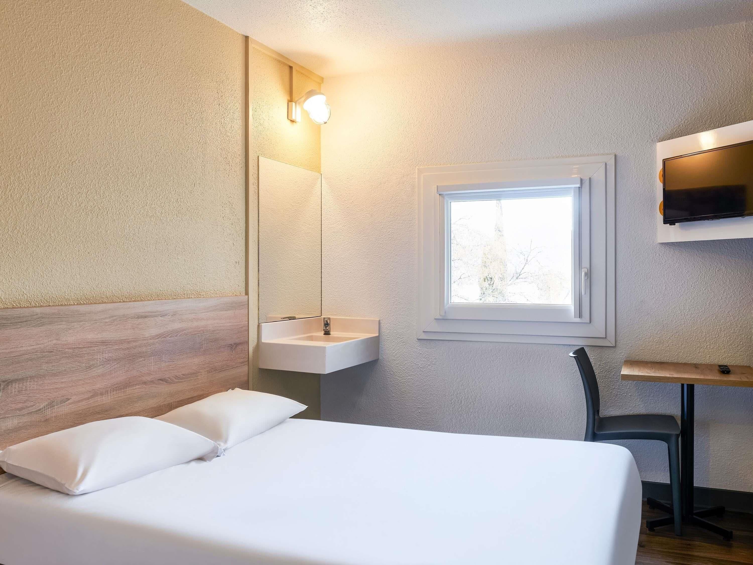 A bedroom with a bed, a bedside table, a mirror, a window, and a sink.