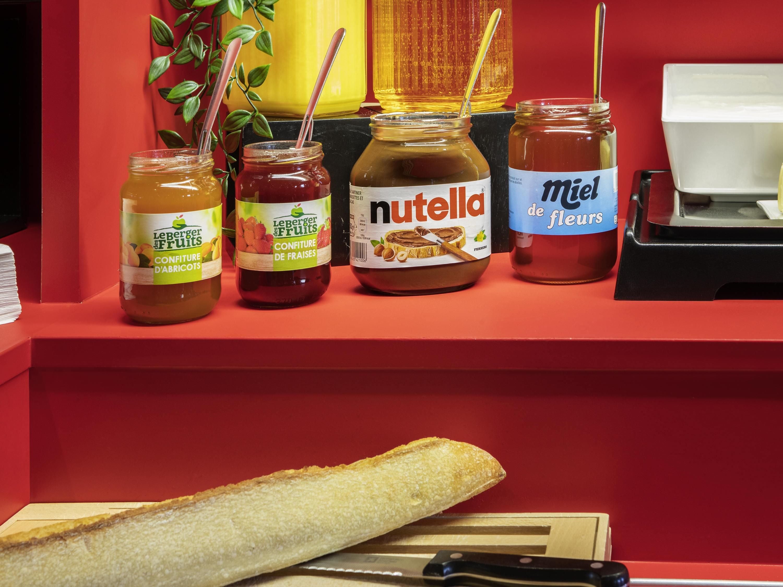 A kitchen counter with jars of Leberger Fruits, Nutella, and Miel de fleurs, and a baguette on a cutting board.