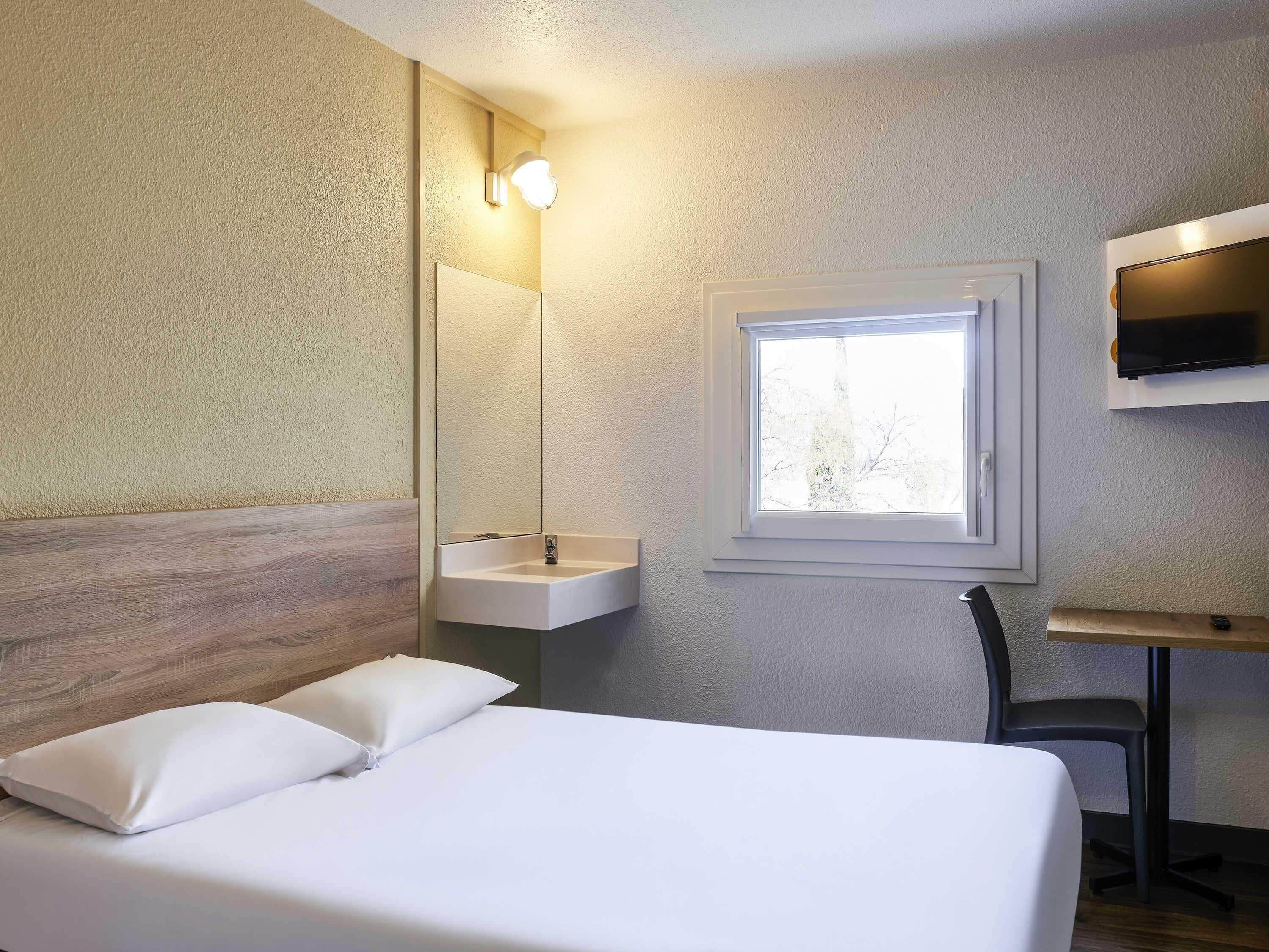 A bedroom with a bed, a bedside table, a mirror, a window, and a wall-mounted TV.
