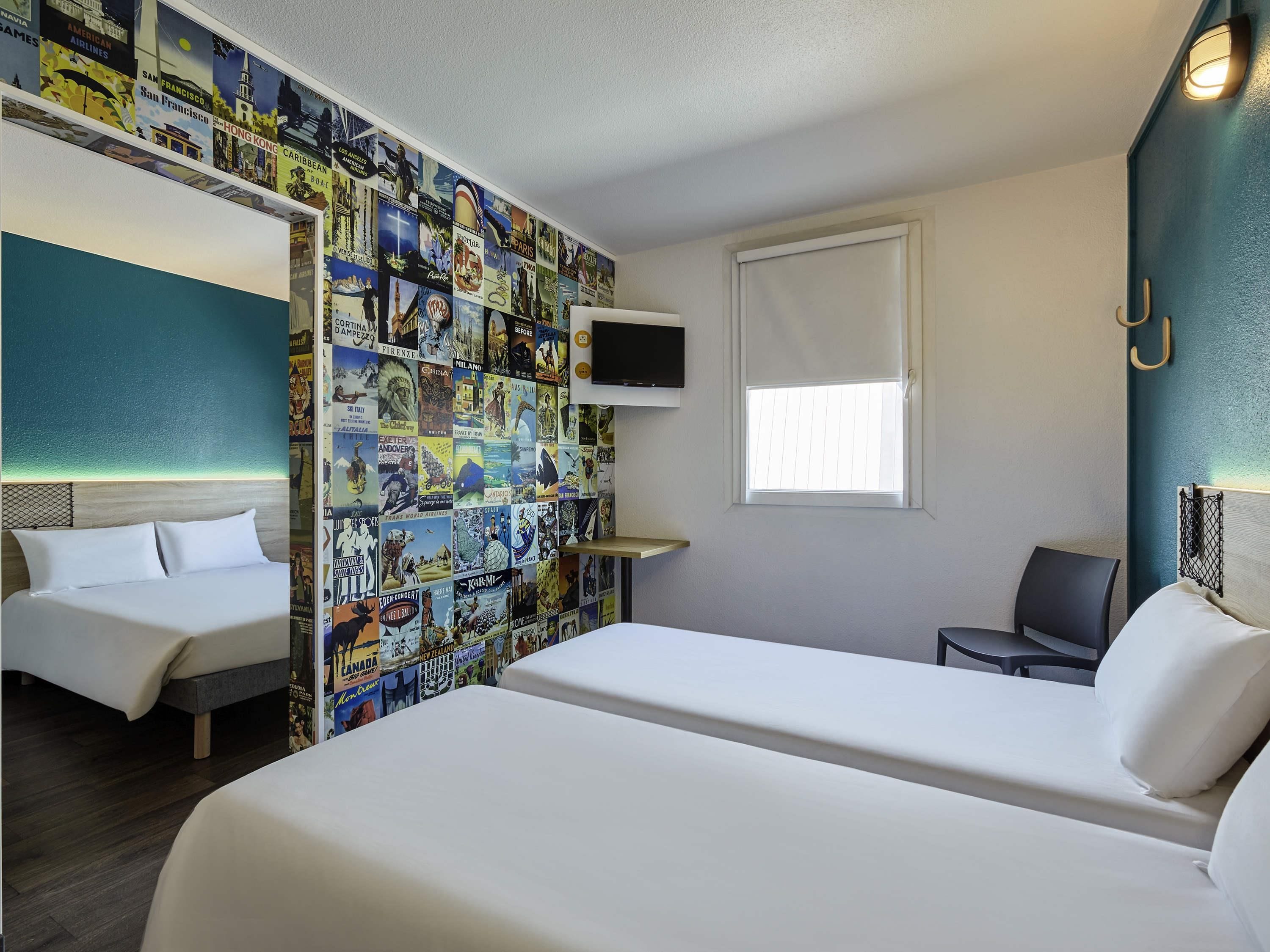 A hotel room with a bed, a TV, a window, and a wall decorated with various travel posters.