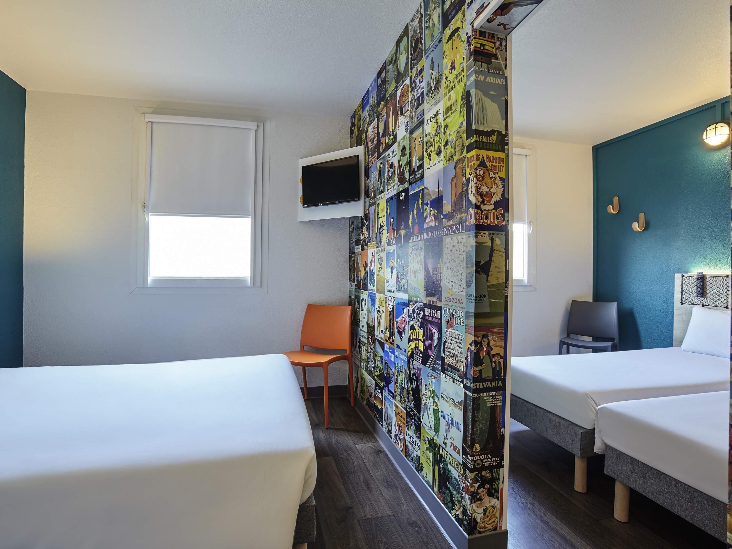 Una camera d'albergo con due letti, una TV fissata al muro, una finestra con persiana bianca e una parete decorata con manifesti di viaggio.