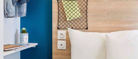 A hotel room with a wooden headboard, a bed with white bedding, a small cactus on a shelf, and a wall-mounted power outlet.
