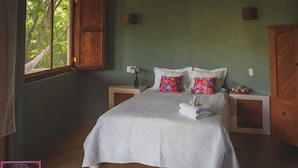 Un dormitorio con una cama grande, dos mesitas de noche, un ventanal con vistas a árboles y un armario de madera.