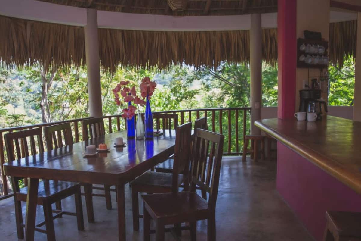 Un comedor con una mesa y sillas de madera, un jarrón azul con flores, y una zona de bar con una estantería llena de vasos.