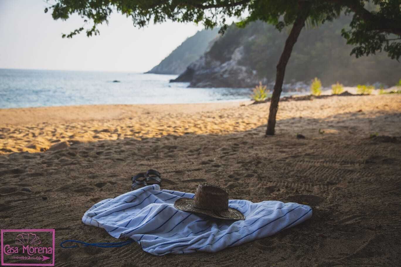 En strandscen med en hatt och en handduk på sanden, ett träd och en klippa i bakgrunden.