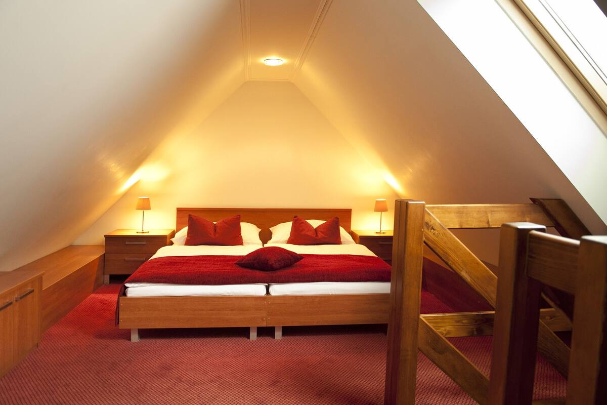 A bedroom with a sloped ceiling, a wooden bed frame, red bedding, and two bedside lamps.