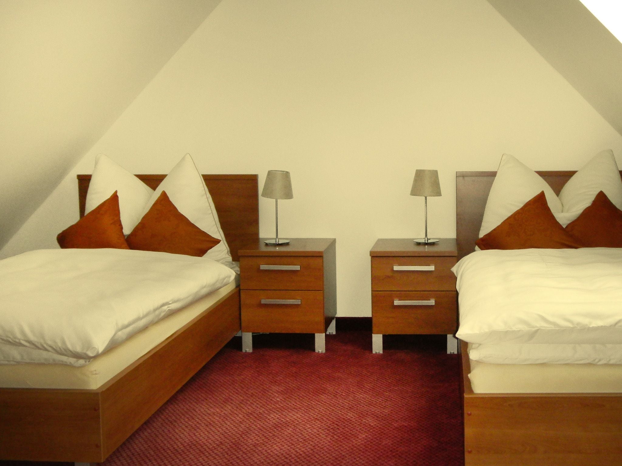 A bedroom with two single beds, each with a wooden headboard, white bedding, and brown pillows. There are two bedside tables with lamps and drawers. The room has a red carpet and a sloped ceiling.