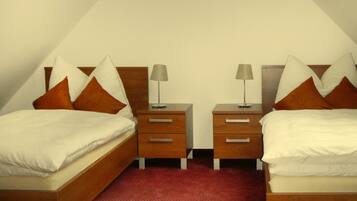 A bedroom with two single beds, each with a wooden headboard, white bedding, and brown pillows. There are two bedside tables with lamps and drawers. The room has a red carpet and a sloped ceiling.