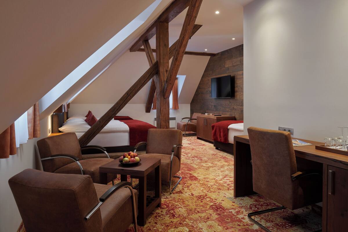A modern hotel room with a sloped ceiling, wooden beams, a flat-screen TV, a desk with a chair, and two beds with red bedding.