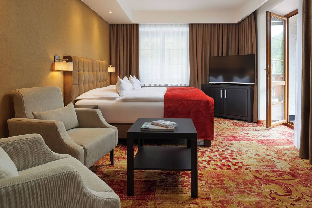 A hotel room with a bed, two armchairs, a small table, a TV, and a large window with curtains.