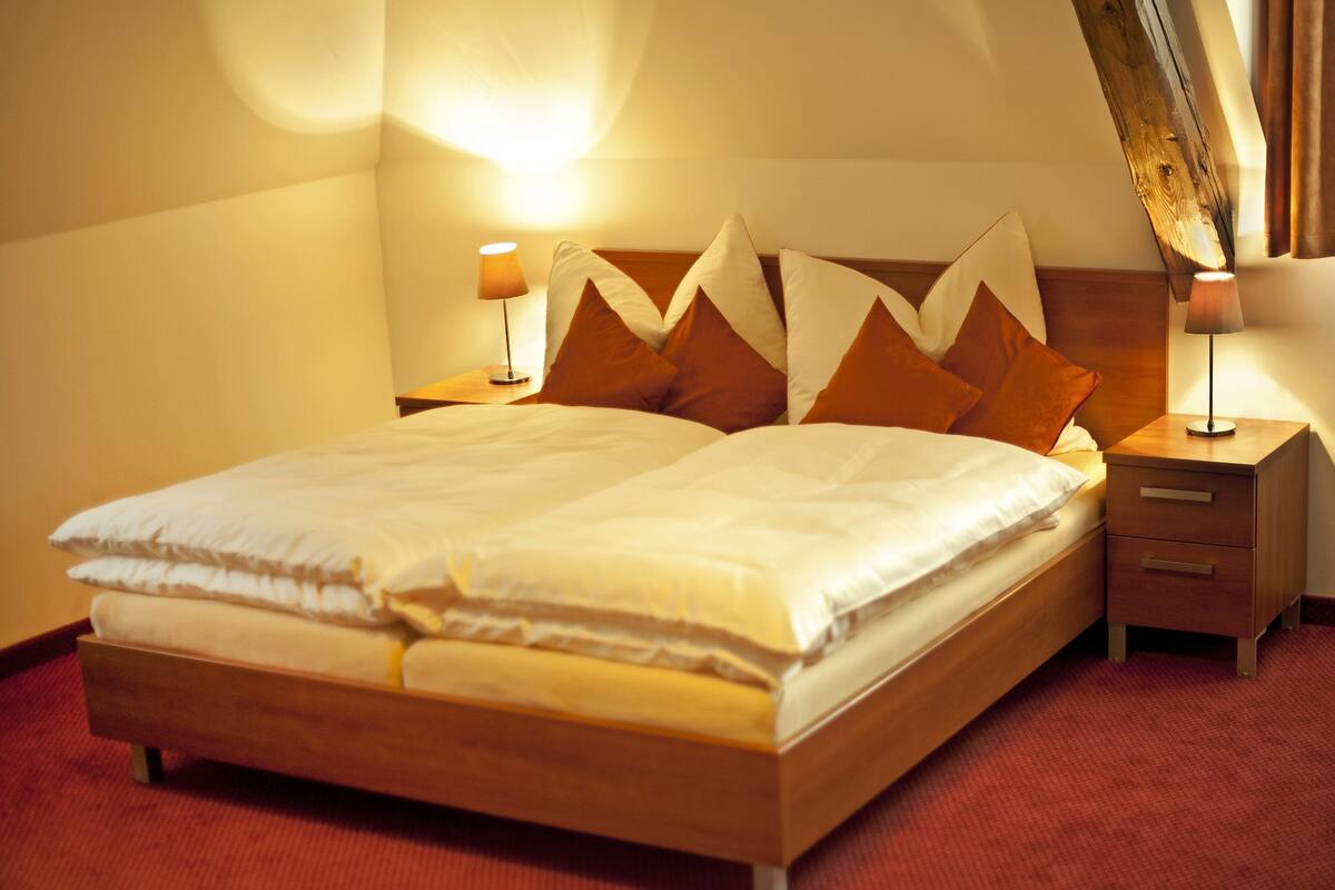 A neatly made bed with a wooden frame and a red carpeted floor. There are two bedside tables with lamps on either side of the bed.