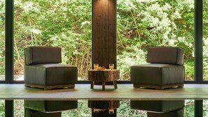 Une pièce moderne avec deux fauteuils en cuir, une table en bois sur laquelle sont disposés des fruits et des boissons, et une grande fenêtre donnant sur une forêt.