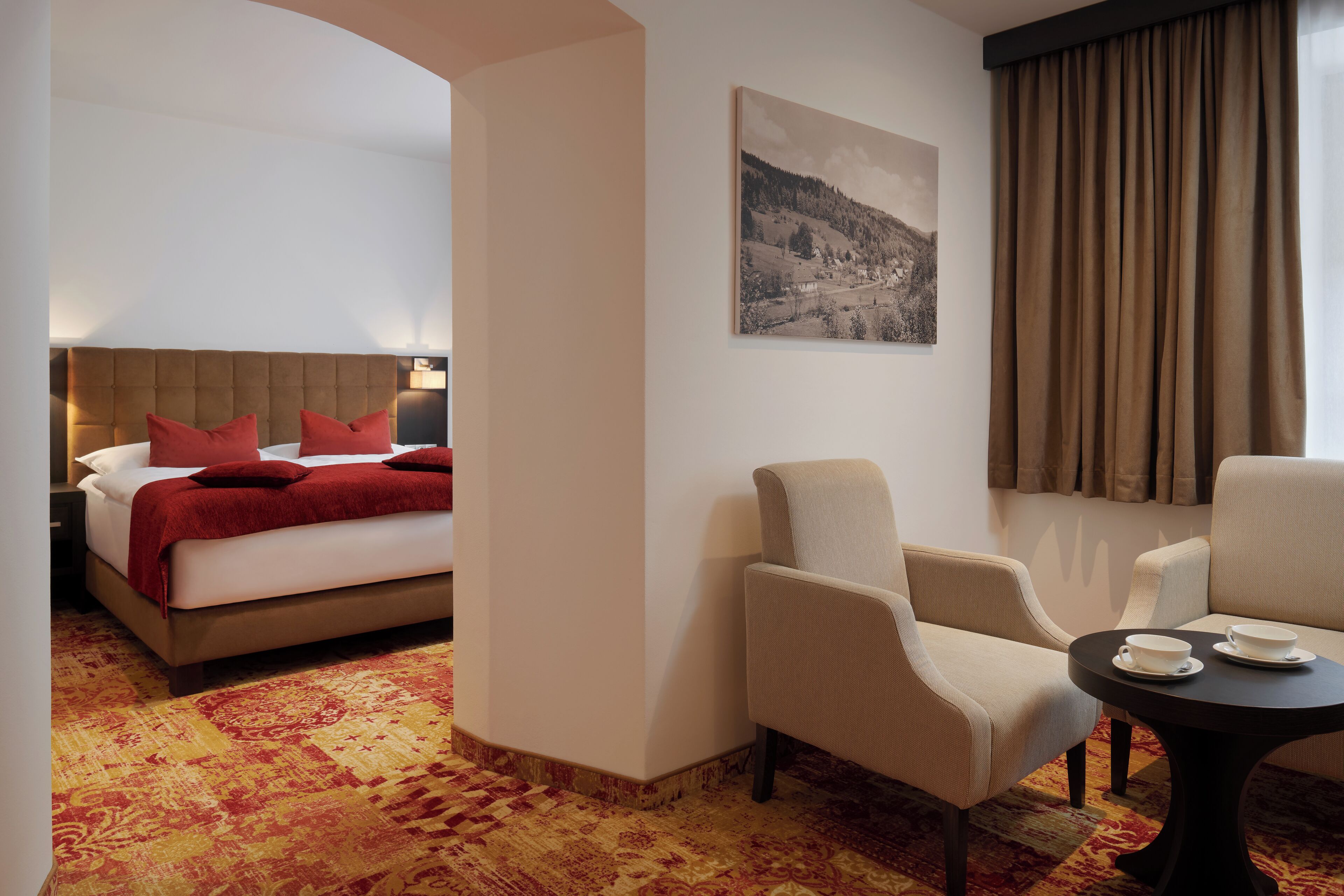 A hotel room with a bed, two armchairs, a small table with cups, a painting on the wall, and a view of a landscape through the window.