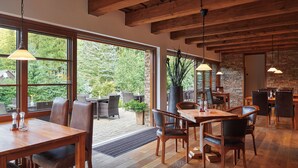 Un vaste espace dédié à la salle à manger, avec des tables et des chaises en bois, de grandes fenêtres donnant sur l’extérieur et un mur en pierre en arrière-plan.