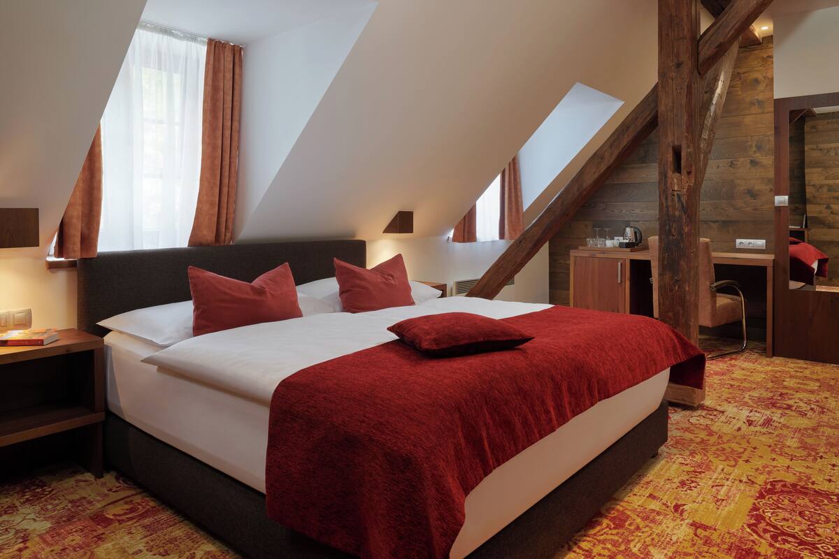 A bedroom with a large bed, a desk, and a chair, featuring a sloped ceiling and wooden beams.