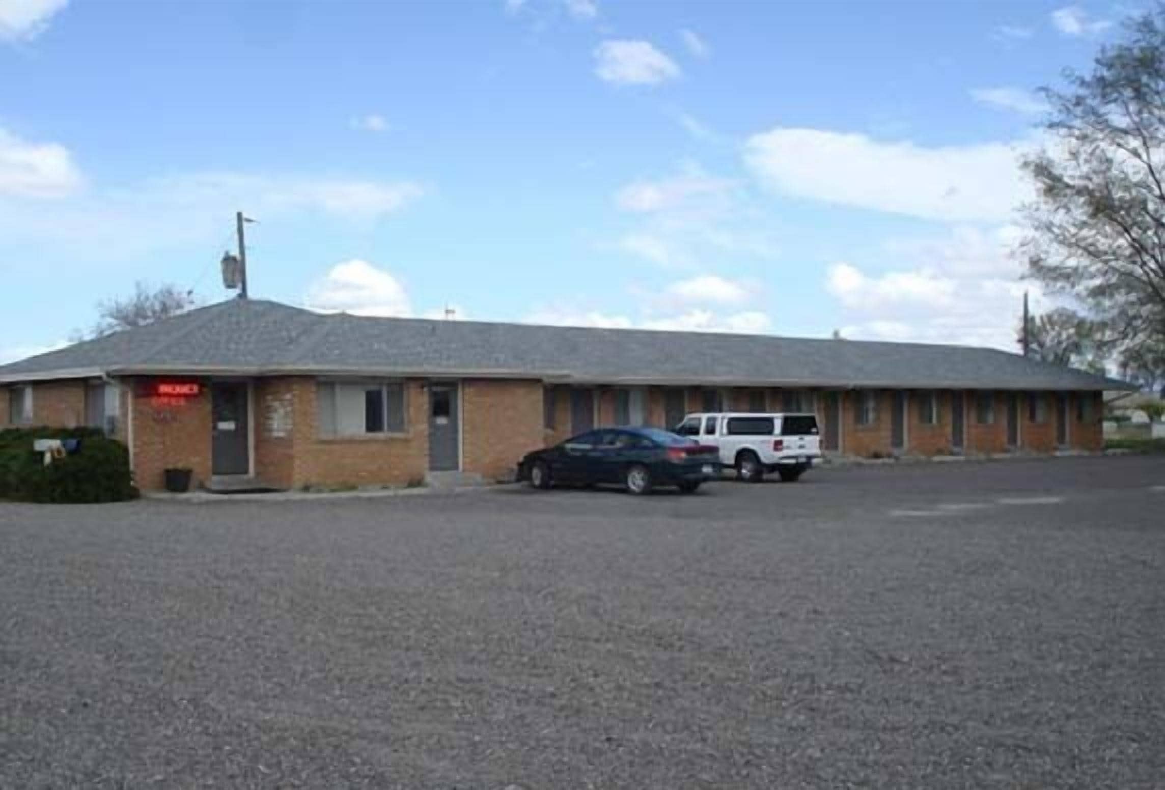 Un motel de una sola planta con fachada de ladrillo, un cartel visible y un estacionamiento con dos autos.