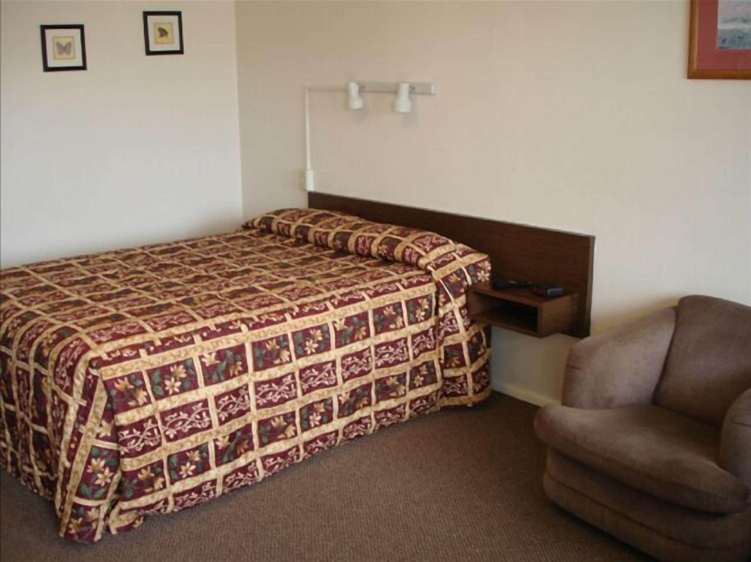 Una cama individual con colcha estampada, cabecera de madera, mesita de noche con lámpara y sillón marrón.