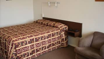 Una cama individual con colcha estampada, cabecera de madera, mesita de noche con lámpara y sillón marrón.