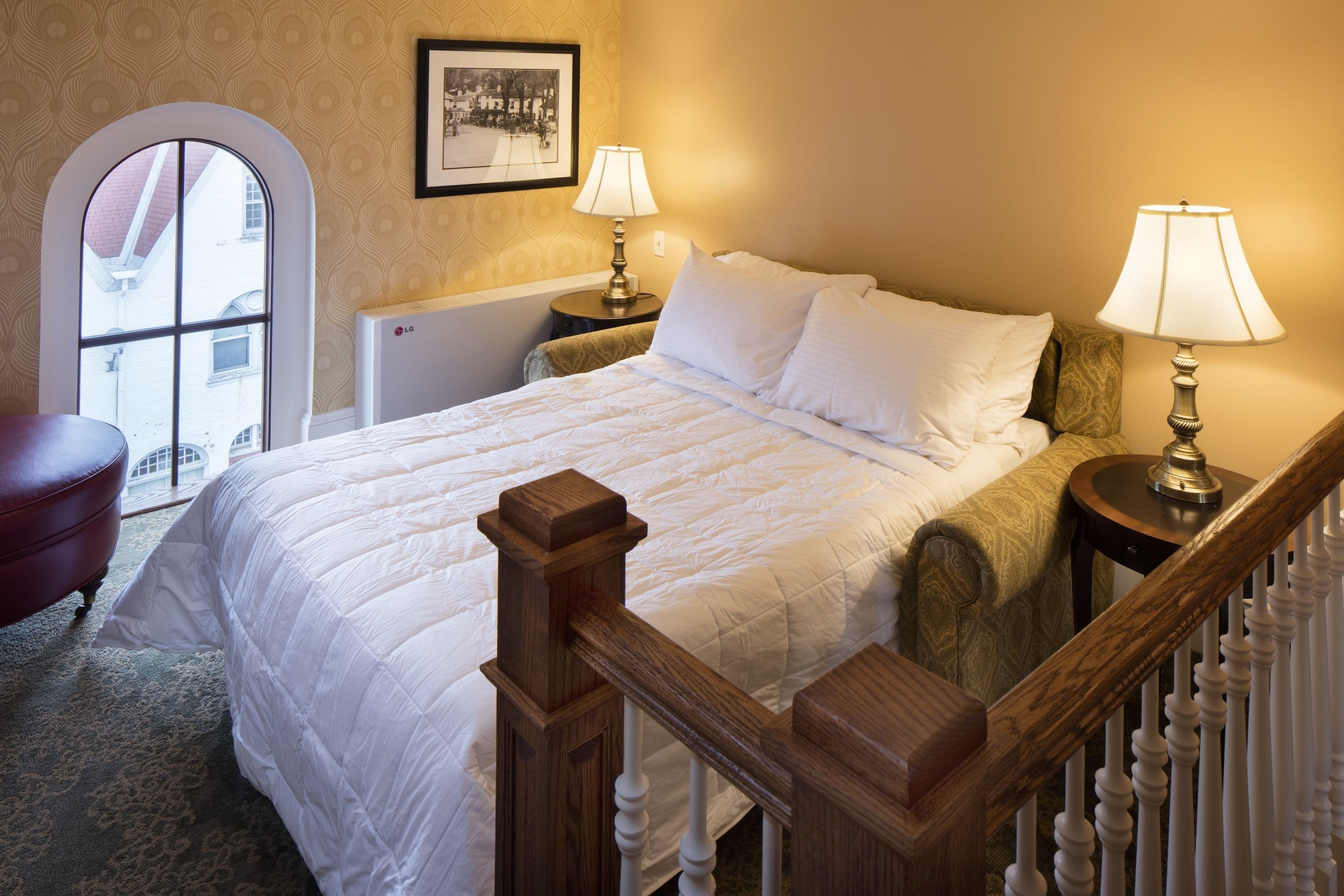 A neatly made bed with white linens, two bedside lamps, a wooden headboard, and a view through an arched window.