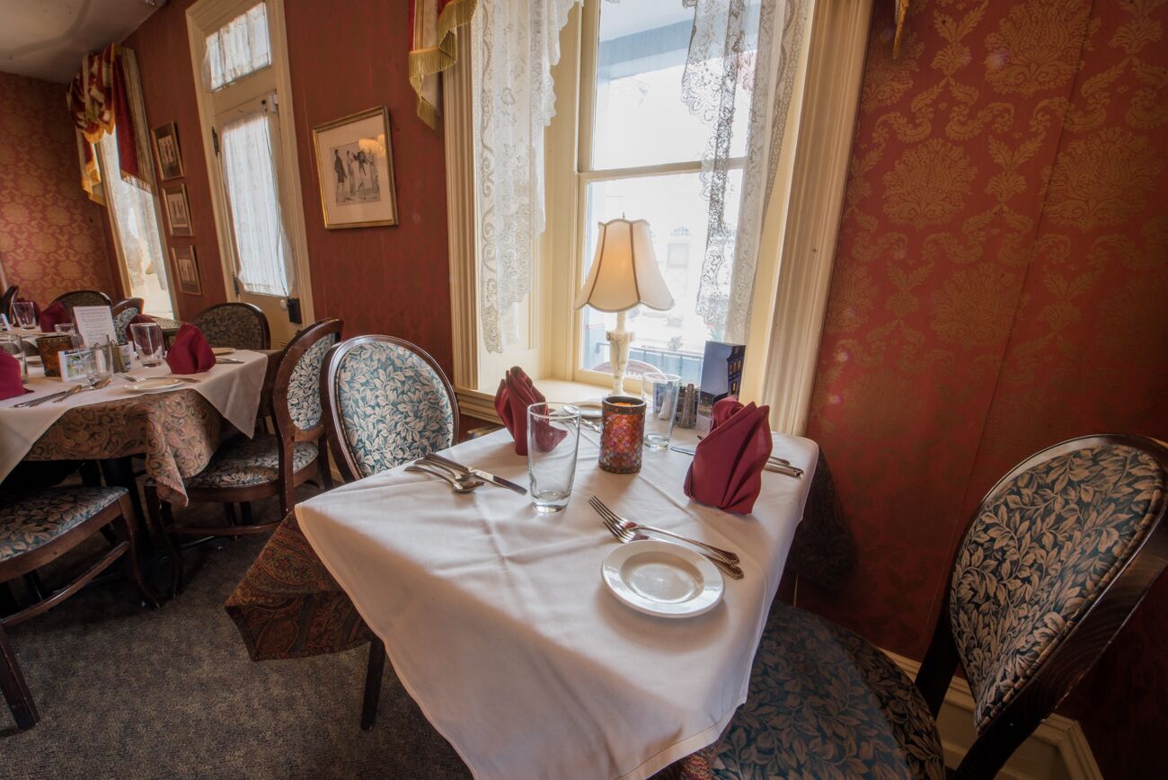 A dining area with a table set for two, featuring a white tablecloth, two chairs with floral upholstery, a candle, and a window with lace curtains.